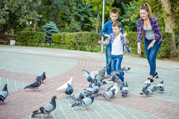 Young beautiful mother and her two sons enjoying together fed pigeons. Happy family. Positive human emotions, feelings, joy. - Фото, изображение