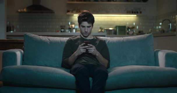 νεαρός άνδρας κάθεται σε έναν καναπέ που χρησιμοποιεί το κινητό του τηλέφωνο δεν δίνοντας προσοχή στην τηλεόραση. - Πλάνα, βίντεο