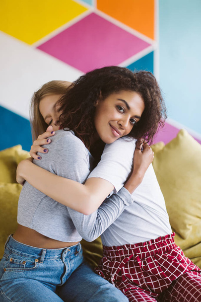 Young smiling african american woman with dark curly hair hugging girlfriend dreamily looking in camera with colorful wall on background  - Foto, Bild