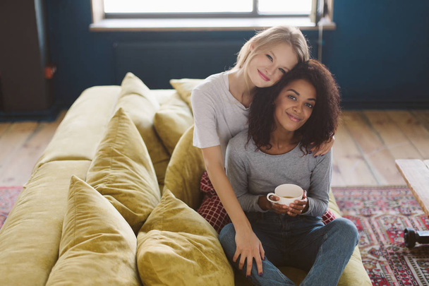 Jonge mooie Afrikaanse Amerikaanse meisje met donkere krullend haar en lachende meisje met blond haar, gelukkig op zoek tijd in de camera doorbrengen samen thuis - Foto, afbeelding