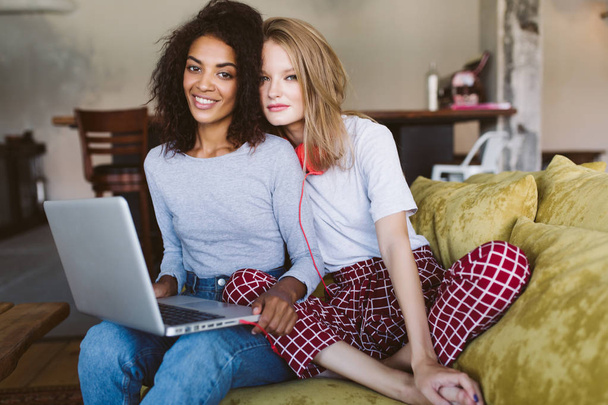 Jeune femme afro-américaine souriante aux cheveux bouclés foncés et ordinateur portable sur les genoux et belle femme aux cheveux blonds et écouteurs rouges regardant à la caméra ensemble à la maison
 - Photo, image