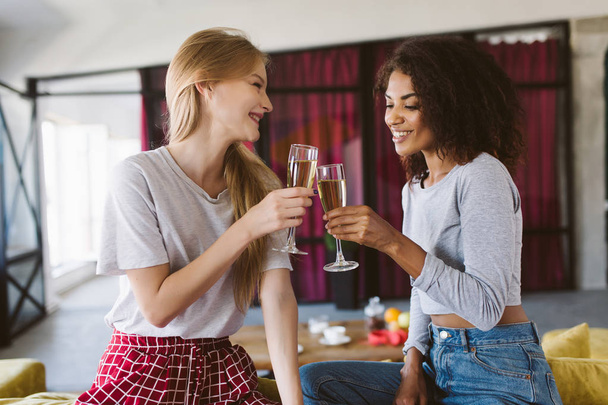 Attractive african american woman with dark curly hair and smiling woman with blond hair sitting on sofa holding glasses of champagne in hands joyfully spending time together at home - Photo, Image