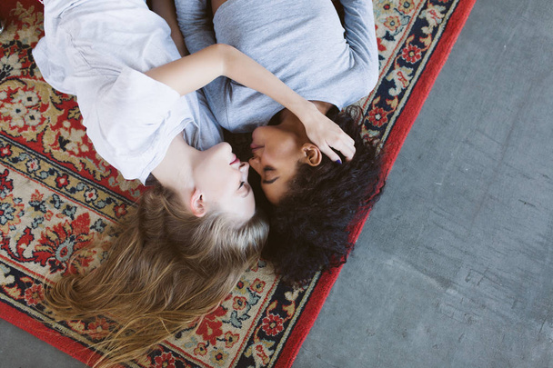 Young beautiful woman with blond hair and african american woman with dark curly hair dreamily lying on vintage carpet spending time together at cozy home - Foto, afbeelding