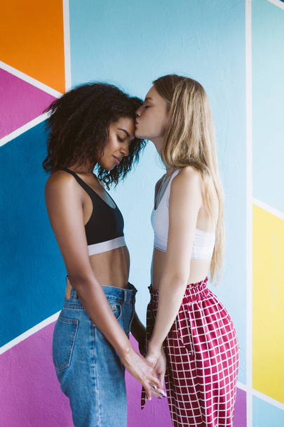 Young beautiful woman with blond hair dreamily kissing forehead of pretty african american woman with dark curly hair holding hands of each other with colorful wall on background - Photo, Image
