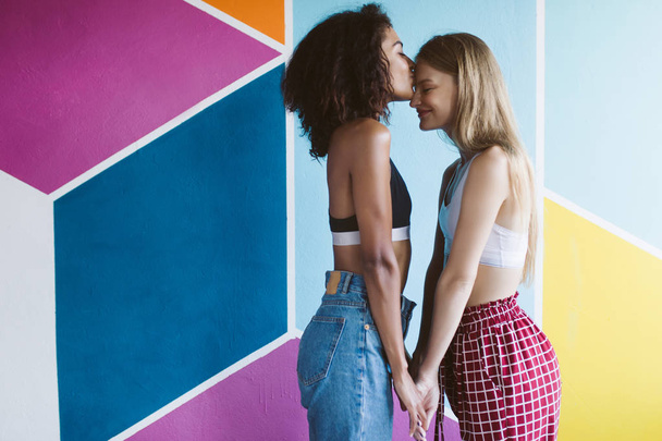 Young nice african american woman with dark curly hair happily kissing forehead of pretty woman with blond hair holding hands of each other with colorful wall on background - Photo, Image