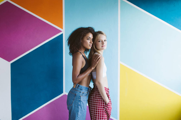 Young nice african american woman with dark curly hair and pretty woman with blond hair in sporty tops dreamily looking aside together with colorful wall on background - Photo, Image