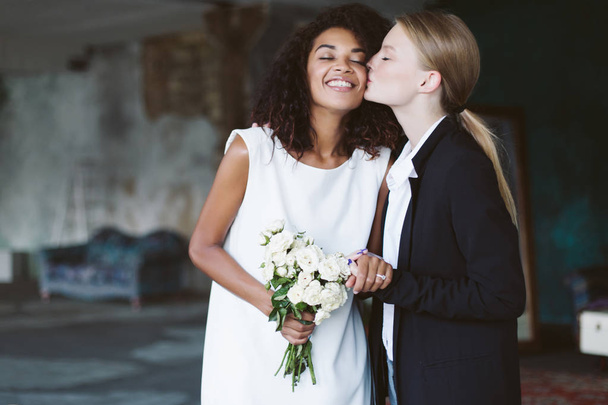 Young woman with blond hair in black suit kissing in cheek pretty african american woman with dark curly hair in white dress with bouquet of flowers in hand on wedding ceremony - Fotó, kép
