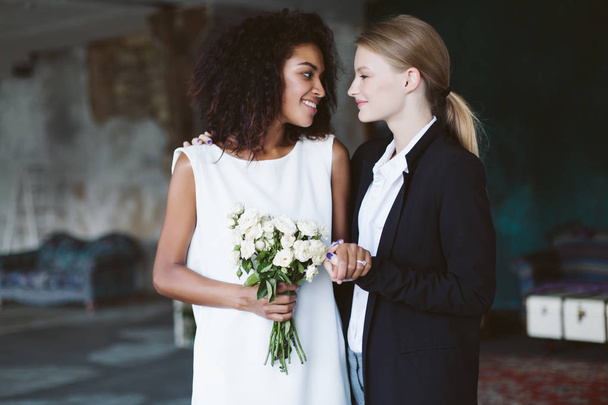 Młoda kobieta z blond włosy w czarnym garniturze i uśmiechający się african american Kobieta, ciemne kręcone włosy w białej sukni z bukietem kwiatów w ręku szczęśliwie patrząc na siebie nawzajem na ślub - Zdjęcie, obraz