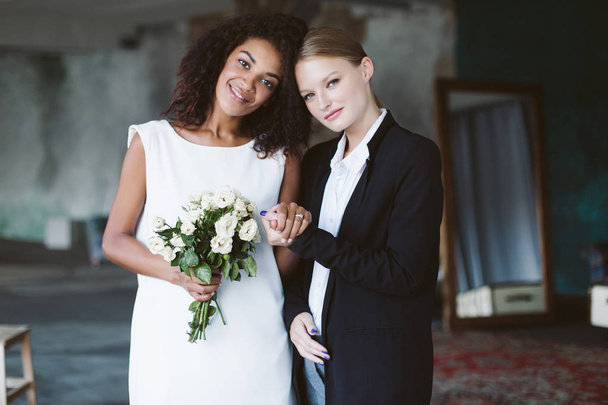 若いきれいな黒のスーツでブロンドの髪を持つ女性、美人喜んでカメラで見ている結婚式の花の小さな花束と白いドレスの暗い巻き毛を持つアフリカ系アメリカ人 - 写真・画像