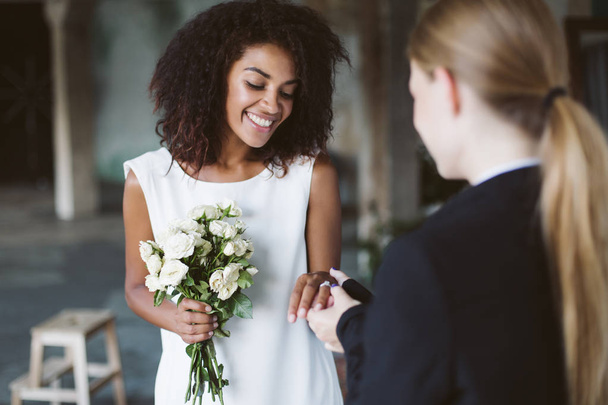 Beautiful african american woman with dark curly hair in white dress holding little bouquet of flowers in hand happily spending time on wedding ceremony - Foto, immagini
