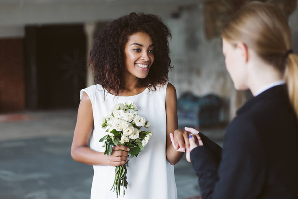 junge attraktive afrikanisch-amerikanische Frau mit dunklem lockigem Haar in weißem Kleid mit kleinem Blumenstrauß in der Hand, die freudig Zeit mit der Hochzeitszeremonie verbringt - Foto, Bild