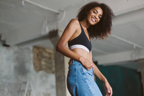 junge hübsche lächelnde Afroamerikanerin mit dunklem lockigem Haar in schwarzem, sportlichem Oberteil und Jeans, die glücklich in die Kamera schaut und Zeit zu Hause verbringt - Foto, Bild