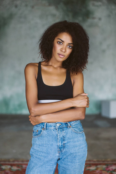 Joven chica afroamericana atractiva con cabello rizado oscuro en top deportivo negro y jeans que miran pensativamente en la cámara pasar tiempo en casa
 - Foto, imagen