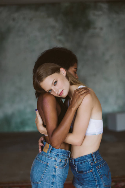Όμορφη γυναίκα με ξανθά μαλλιά στην κορυφή αγκαλιάζει αφρικανική αμερικανική φίλη απορώντας ψάχνει στην κάμερα  - Φωτογραφία, εικόνα