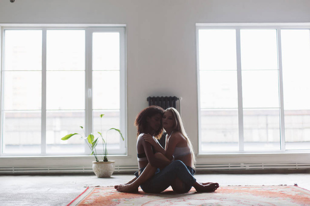 Young attractive woman with blond hair and african american woman with dark curly hair sitting on carpet dreamily looking in camera spending time together at home with big windows on background - Photo, image
