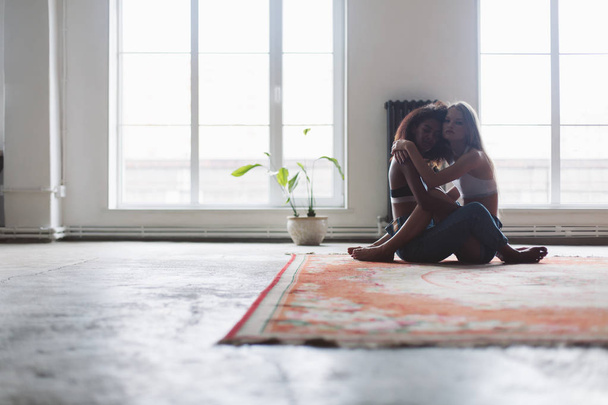Jonge aantrekkelijke vrouw met blond haar en african american vrouw met donker krullend haar zittend op tapijt zorgvuldig in de camera kijkt met grote ramen op de achtergrond - Foto, afbeelding