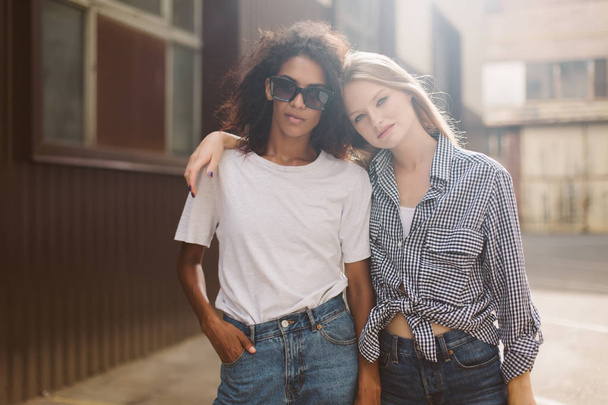 Young african american woman with dark curly hair in sunglasses and T-shirt and pretty woman with blond hair in shirt dreamily looking in camera spending time together - 写真・画像
