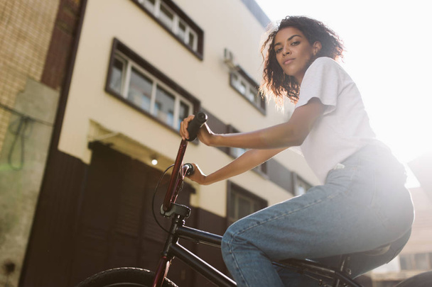junge ernsthafte afrikanisch-amerikanische Frau mit dunklem lockigem Haar in weißem T-Shirt und Jeans, die nachdenklich in die Kamera schaut, während sie Fahrrad fährt - Foto, Bild