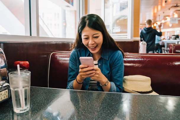 カジュアルな女性がリラックスできる食堂に座って、楽しく食事を待っている間のスマート フォンで彼女の友人とチャットします。若い女性は携帯電話を使用して嬉しそう。ランチ コンセプトを持つアジア. - 写真・画像