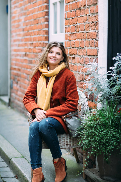 Egy fiatal, szép, szőke nő ül a székre, és élvezi a kis városi utca, Lübeck-ben. Szép l barna kabátot visel. - Fotó, kép