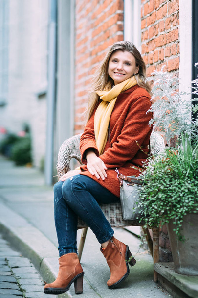 Una joven y hermosa mujer rubia está sentada en la silla y disfrutando de una pequeña calle urbana en Lubeck. Ella lleva un bonito abrigo marrón.
. - Foto, Imagen
