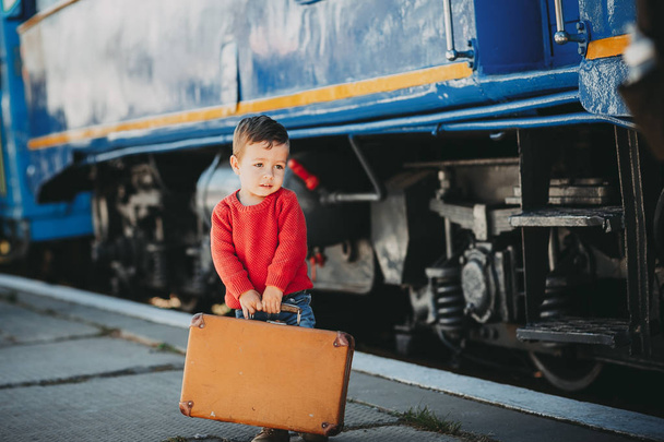 Adorable little kid boy dressed in red sweater on a railway station near train with retro old  brown suitcase. Ready for vacation. Young traveler on the platform. - Φωτογραφία, εικόνα