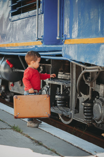 Suloinen pikkupoika punaisessa puserossa juna-asemalla retro vanhan ruskean matkalaukun kanssa. Valmiina lomalle. Nuori matkustaja laiturilla
. - Valokuva, kuva