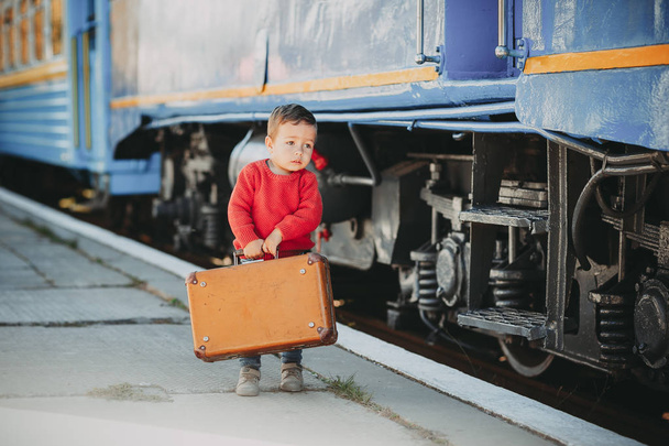 Adorable little kid boy dressed in red sweater on a railway station near train with retro old  brown suitcase. Ready for vacation. Young traveler on the platform. - Photo, Image