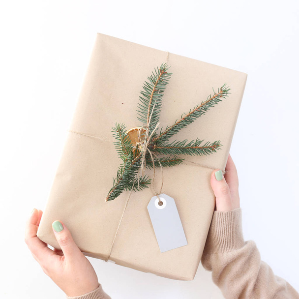 Cadeau de Noël du Nouvel An en papier artisanal décoré d'une branche de sapin avec une étiquette pour signer dans les mains des femmes sur un fond blanc. Vue de dessus, plan plat
 - Photo, image
