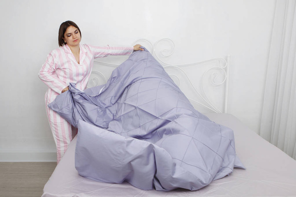 Jonge leuke donkere harige vrouw in roze pyjama maakt het bed met kussens en blauwe beddengoed. - Foto, afbeelding