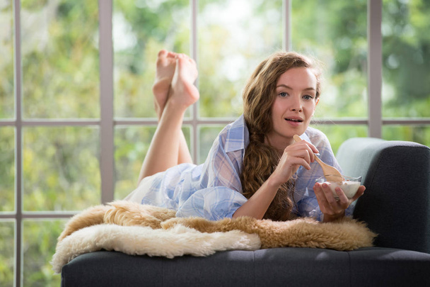 Mulher jovem saudável deitado em um sofá segurando uma tigela de iogurte olhando relaxado e confortável
 - Foto, Imagem