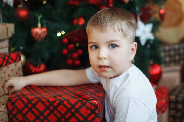 Boy in Christmas hats is sitting with gift boxes in his hands with the joy of a surprise. - Foto, afbeelding