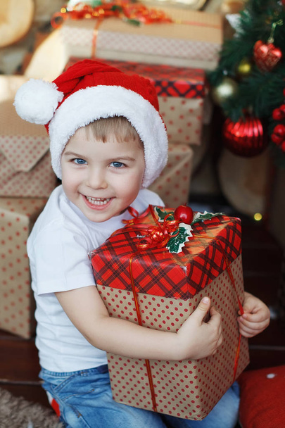 クリスマスの帽子の少年はギフト用の箱と驚きの喜びの彼の手に座っています。. - 写真・画像