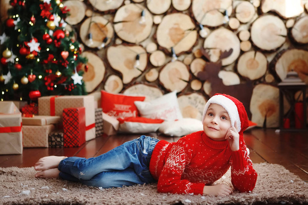 Poika jouluhatut makaa ja nauraa matolla lähellä uudenvuoden puu taustalla puun leikkauksia
. - Valokuva, kuva