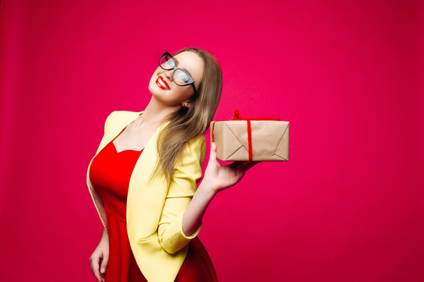 Beautiful happy woman in fashionable cat-eye glasses in bright dress holding Christmas present. - Photo, Image