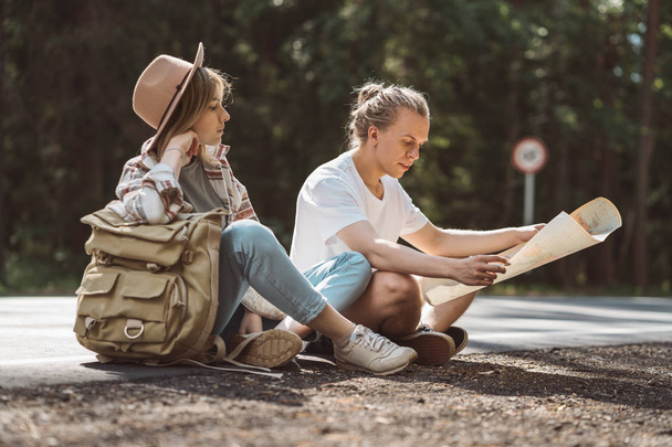 Couple of travelers sitting on the side of the road rest and look at the location map. Travelers hipsters woman and man with backpack hold in hands location map and searching directional - Photo, Image