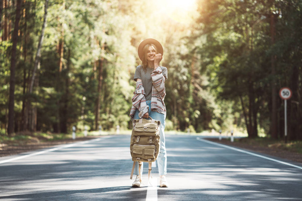 Young happy woman with traveling backpack wearing hat stand alone on countryroad. Traveler girl walking along the road to forest - Foto, immagini