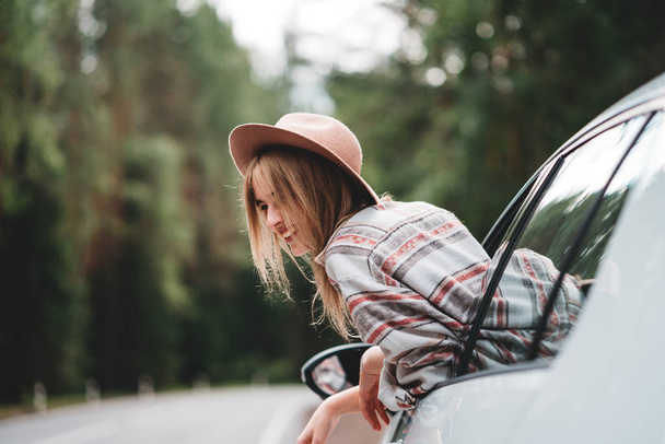 Handsome woman hanging out of the car window, hold vintage hat and enjoying beautiful  road in forest - Zdjęcie, obraz