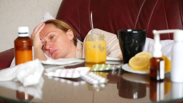 sairas nainen makaa olohuoneensa sohvalla, hänen vieressään pöydällä on lääkkeitä ja kuuma juoma hunajalla ja sitruunalla
. - Valokuva, kuva