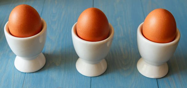 3 вареных яйца в белых стаканах яйца на синем деревянном фоне
 - Фото, изображение