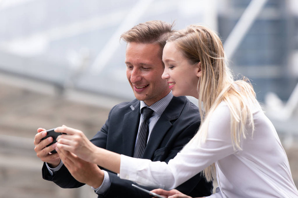 пара деловых людей, смотрящих на смартфон вместе и улыбающихся на фоне офисного здания
. - Фото, изображение