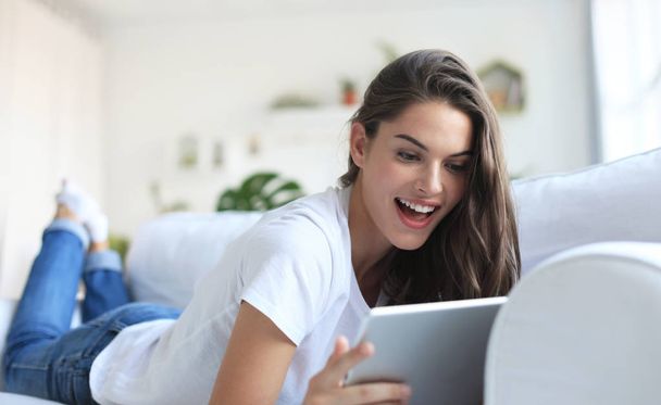 Beautiful young woman using tablet sitting on sofa at home at living room. - 写真・画像