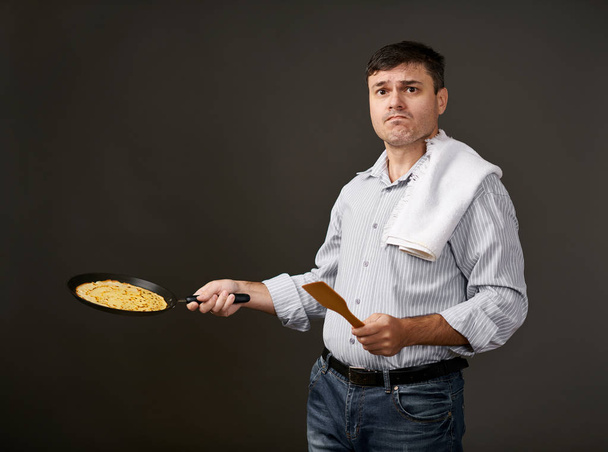Mann posiert mit Pfannkuchen in der Pfanne, weißem Hemd und weißer Hose, grauem Hintergrund, überraschten Emotionen - Foto, Bild