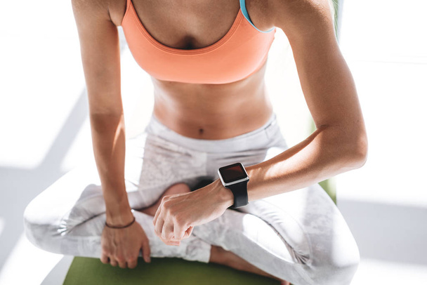 Woman sitting on yoga lotus pose and using smartwatch. Closeup view of young female practicing yoga in training hall - Foto, immagini