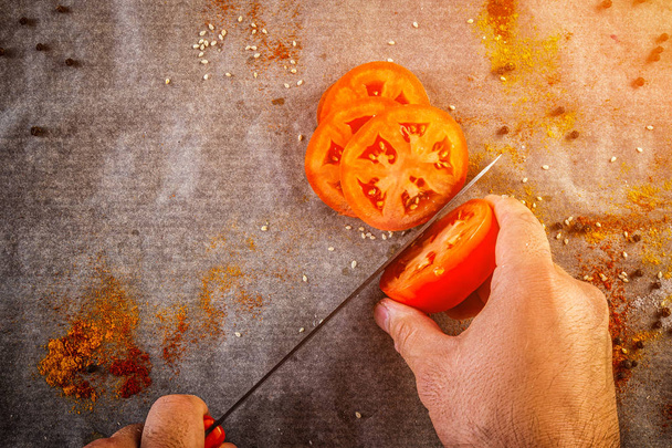 Closeup of male chef hand cutting a ripe red tomato with a kitchen knife against the background of a kitchen table with spices, salt, pepper, top view - Photo, Image