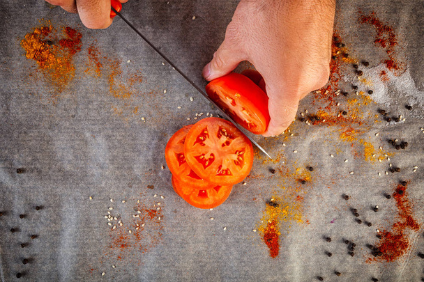 Primer plano del chef masculino cortando a mano un tomate rojo maduro con un cuchillo de cocina contra el fondo de una mesa de cocina con especias, sal, pimienta, vista superior
 - Foto, Imagen