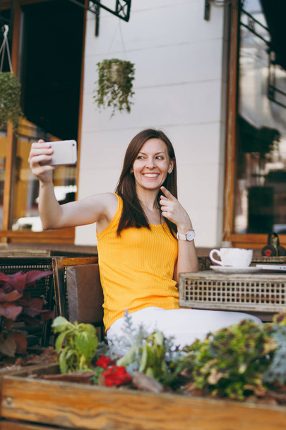 Happy smiling girl in outdoors street coffee shop cafe sitting at table with laptop computer doing taking selfie shot on mobile phone in restaurant during free time. Mobile office freelance concept - Photo, Image