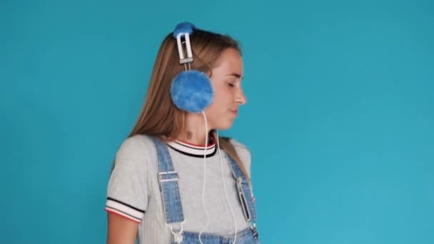Portrait. Cute girl dancing and moving to the rhythm listen to music in headphones. Blue background. Teenage girl wearing headphones - Πλάνα, βίντεο