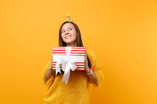 Dreamy young woman in birthday hat looking up, holding red box with gift, present enjoying holiday isolated on bright yellow background. People sincere emotions, lifestyle concept. Advertising area - Foto, immagini