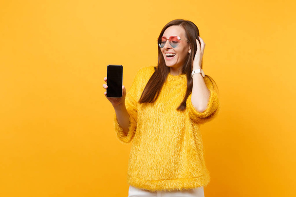 Laughing young woman in heart glasses putting hand on head holding mobile phone with blank black empty screen isolated on bright yellow background. People sincere emotions lifestyle. Advertising area - Foto, Imagen
